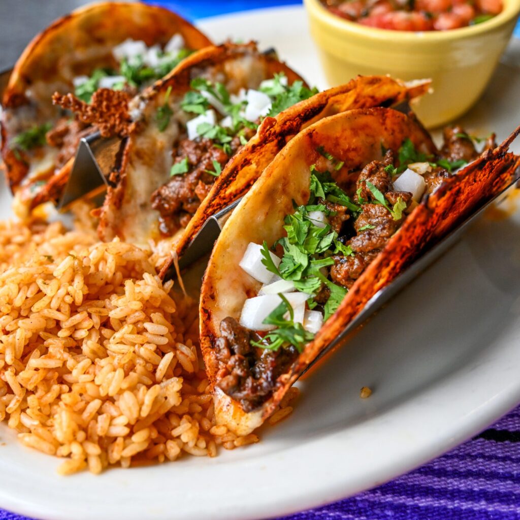 Three white corn tortillas dipped in Birria sauce and filled with Birria steak and melted jack cheese. Topped with raw onions and cilantro. Served with rice, borracho beans, limes and Birria sauce for dipping